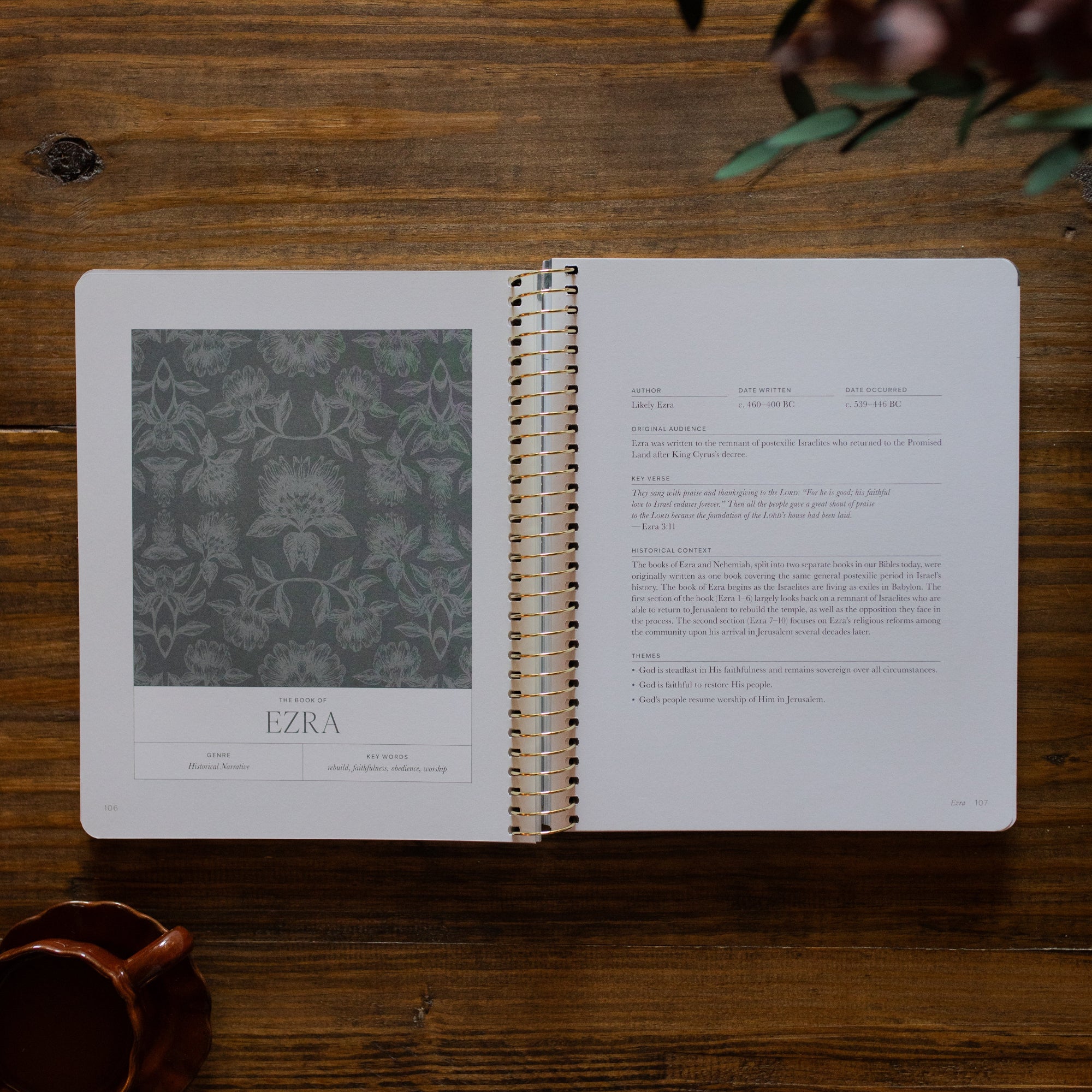 A Year in the Bible Spiral opened to the the Ezra section next to a brown coffee cup on a brown wooden table