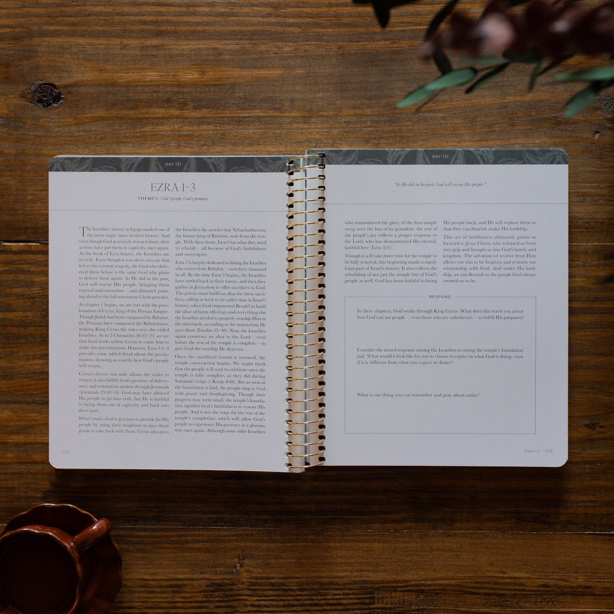 A Year in the Bible Spiral opened to the Ezra reading portion and questions page next to a brown coffee cup on a brown wooden table