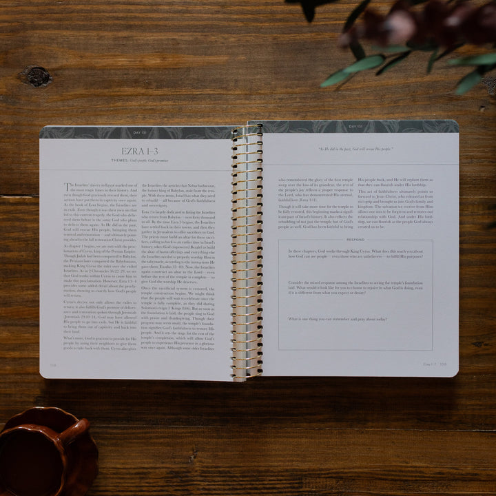 A Year in the Bible Spiral opened to the Ezra reading portion and questions page next to a brown coffee cup on a brown wooden table