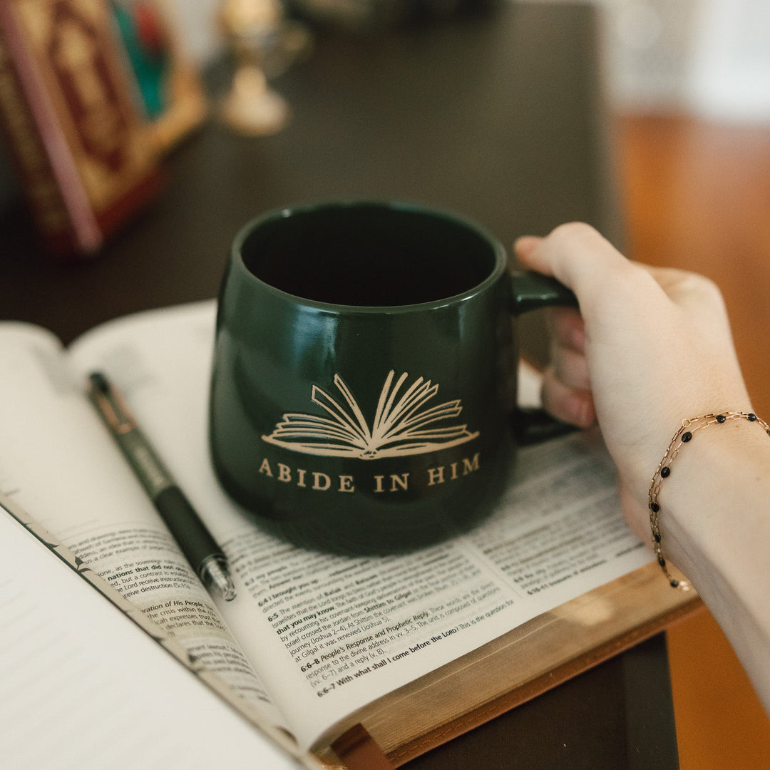 A dark green ceramic mug with a gold book graphic and the words 'ABIDE IN HIM' is held by a hand over an open book on a wooden table.