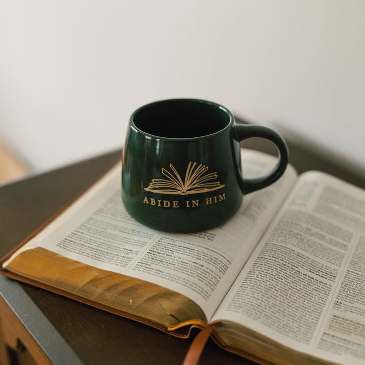 A dark green ceramic mug with a gold book-and-cross design and the words 'ABIDE IN HIM' printed on the side, resting on an open book on a wooden table.
