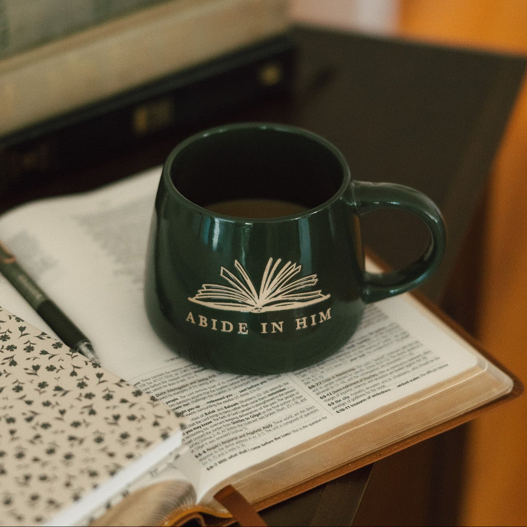A dark green ceramic mug with a white book-and-ribbon graphic and the words 'ABIDE IN HIM' sits on an open book on a table, surrounded by stacked books and a notebook in a warm indoor setting.