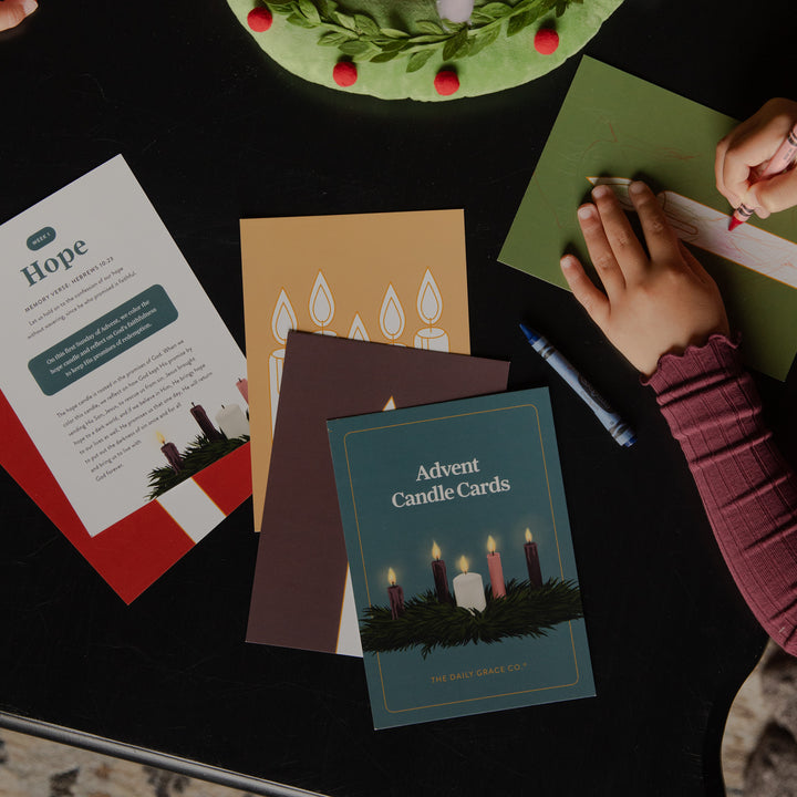 A styled flat-lay of Advent candle cards and writing materials on a black tabletop. Visible items include a teal card set labeled 'Advent Candle Cards,' a white card with 'Hope' text, a gold card with candle illustrations, a green writing pad being held by a child's hand, a blue marker, and part of a green wreath at the top.