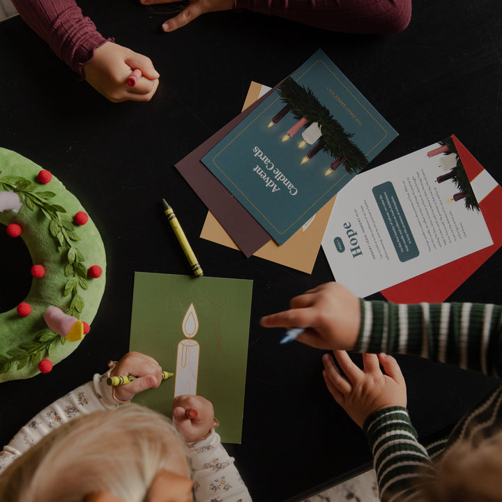 Overhead view of children's hands and heads working on a craft activity at a dark table, with greeting cards, a green wreath, crayons, and paper visible.