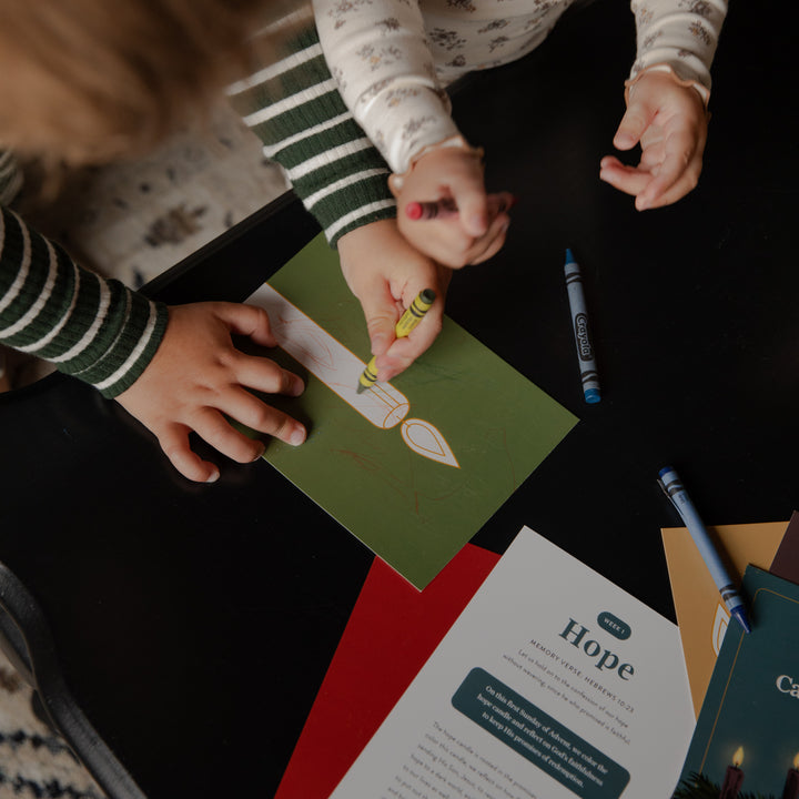 A top-down view of children's hands creating a candle-shaped craft on a green card over a black table, with crayons and printed cards nearby.