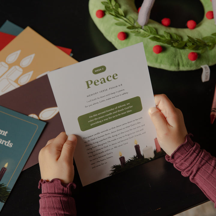 A person holds a printed weekly devotional card titled 'Peace' over a dark tabletop, surrounded by holiday cards, a green wreath with red pom-poms, and candles.