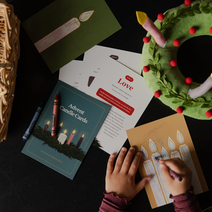 A child's hands are shown writing on an advent candle card, surrounded by advent-themed craft supplies including a green felt wreath with red berries, crayons, and printed cards on a dark tabletop.