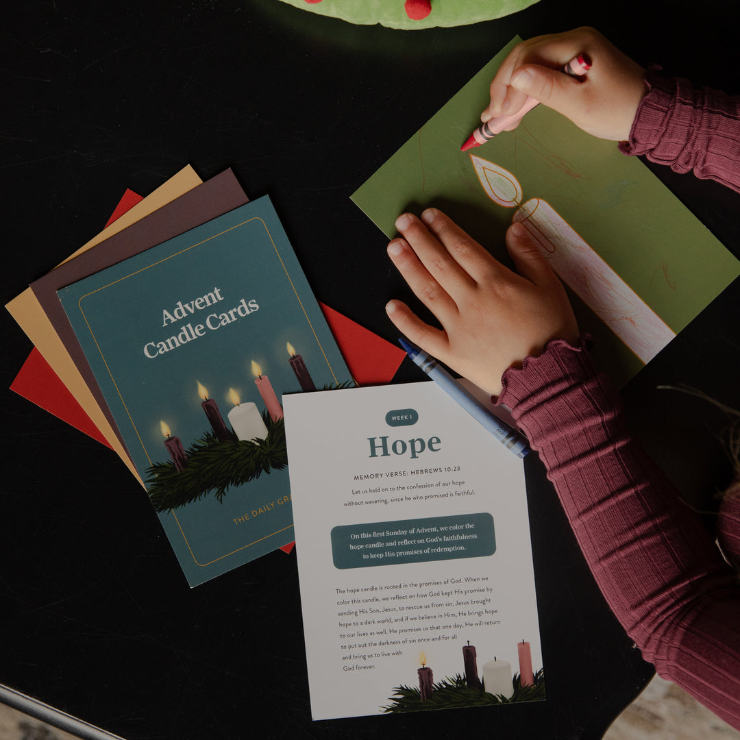 A top-down view of a person's hands creating an Advent candle card set on a dark table. One hand holds a red marker over a green card, while the other rests on a printed instruction sheet titled 'Hope'.