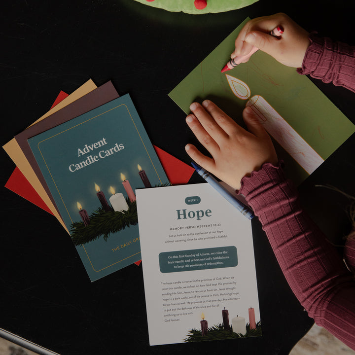 A top-down view of a person's hands creating an Advent candle card set on a dark table. One hand holds a red marker over a green card, while the other rests on a printed instruction sheet titled 'Hope'.