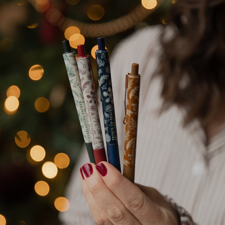 A close-up of a hand holding four decorative ballpoint pens with floral patterns, set against a warm, blurred Christmas tree background with glowing lights.