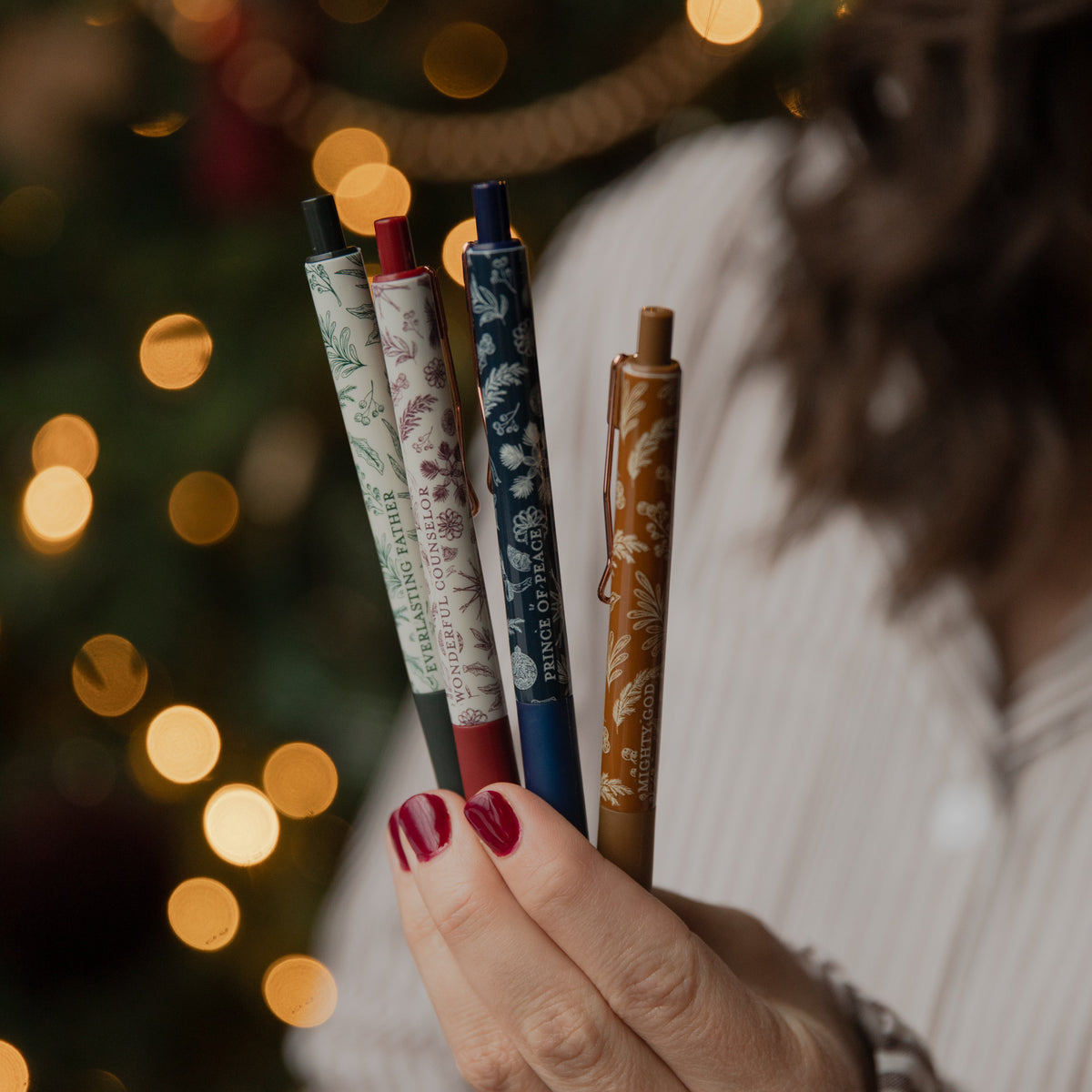 A close-up of a hand holding four decorative ballpoint pens with floral patterns, set against a warm, blurred Christmas tree background with glowing lights.