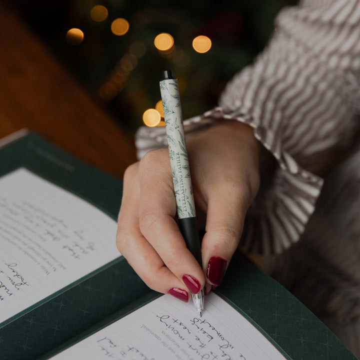 A close-up of a person's hand with red manicured nails writing in a notebook using a pen with a green and white marbled pattern and black accents, set against a warm, blurred background with holiday lights.