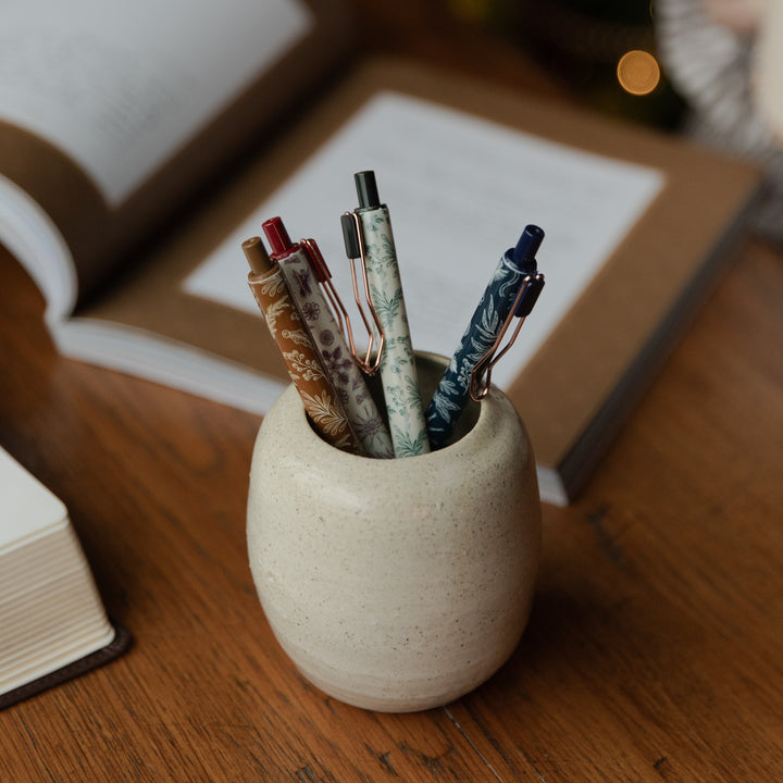 A small round speckled ceramic pen holder on a wooden desk, holding several pens, with blurred books in the background.