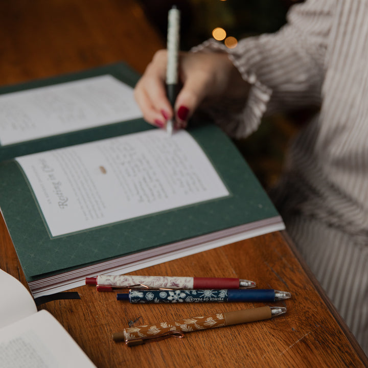 A close-up lifestyle shot of a person writing in a dark green journal with a black pen, with several decorative pens resting on a wooden desk in the foreground.