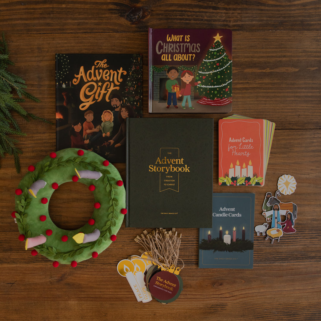 A flat lay of Advent-themed stationery and gift items arranged on a wooden tabletop, including a dark green Advent Storybook, an Advent Gift book, a Christmas card, a wreath with colorful ornaments, a set of Advent Cards for Little Theaters, and small pins.