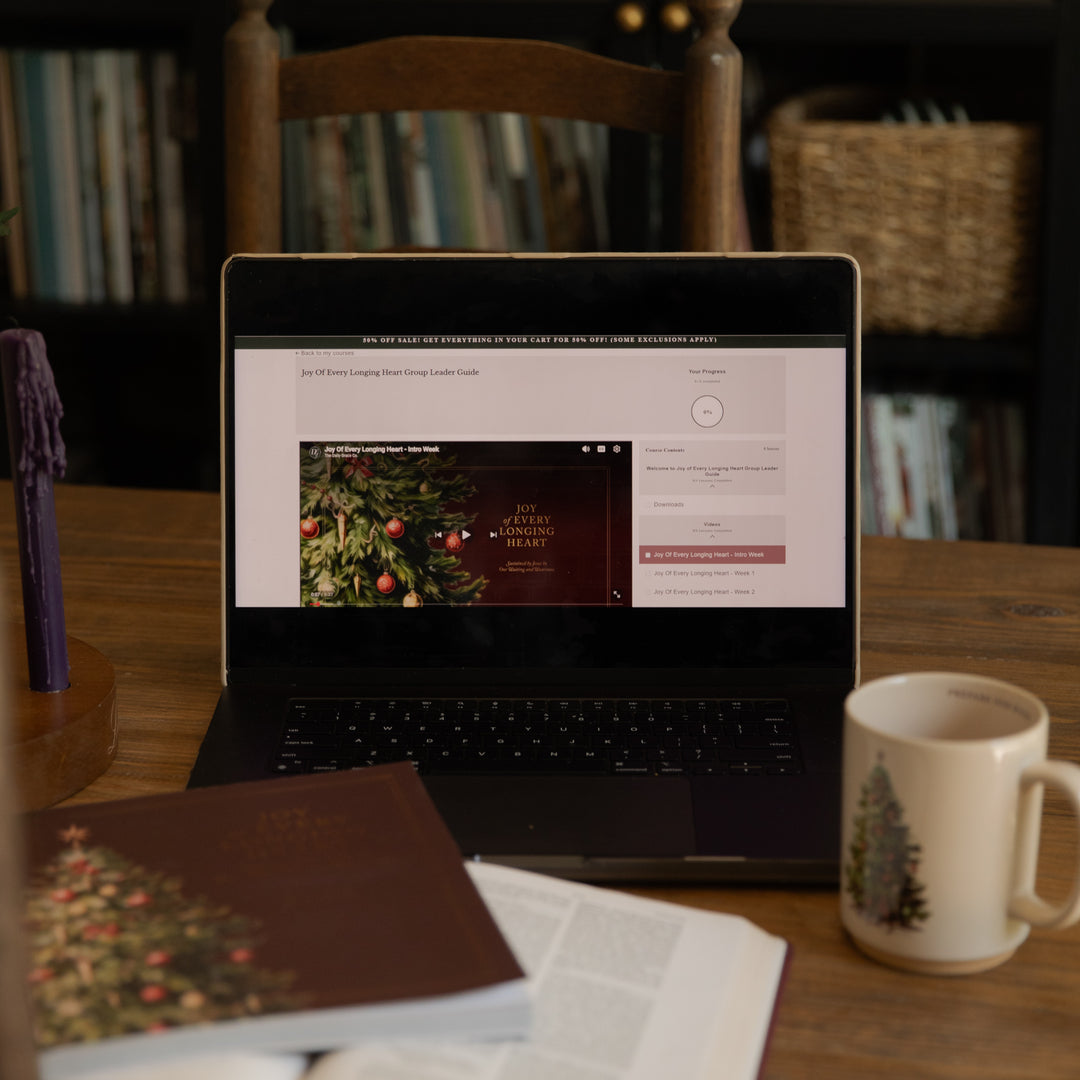 A laptop on a wooden table displaying a website, surrounded by a book, an open magazine, a white mug with a Christmas tree design, and a candle in a cozy indoor setting.