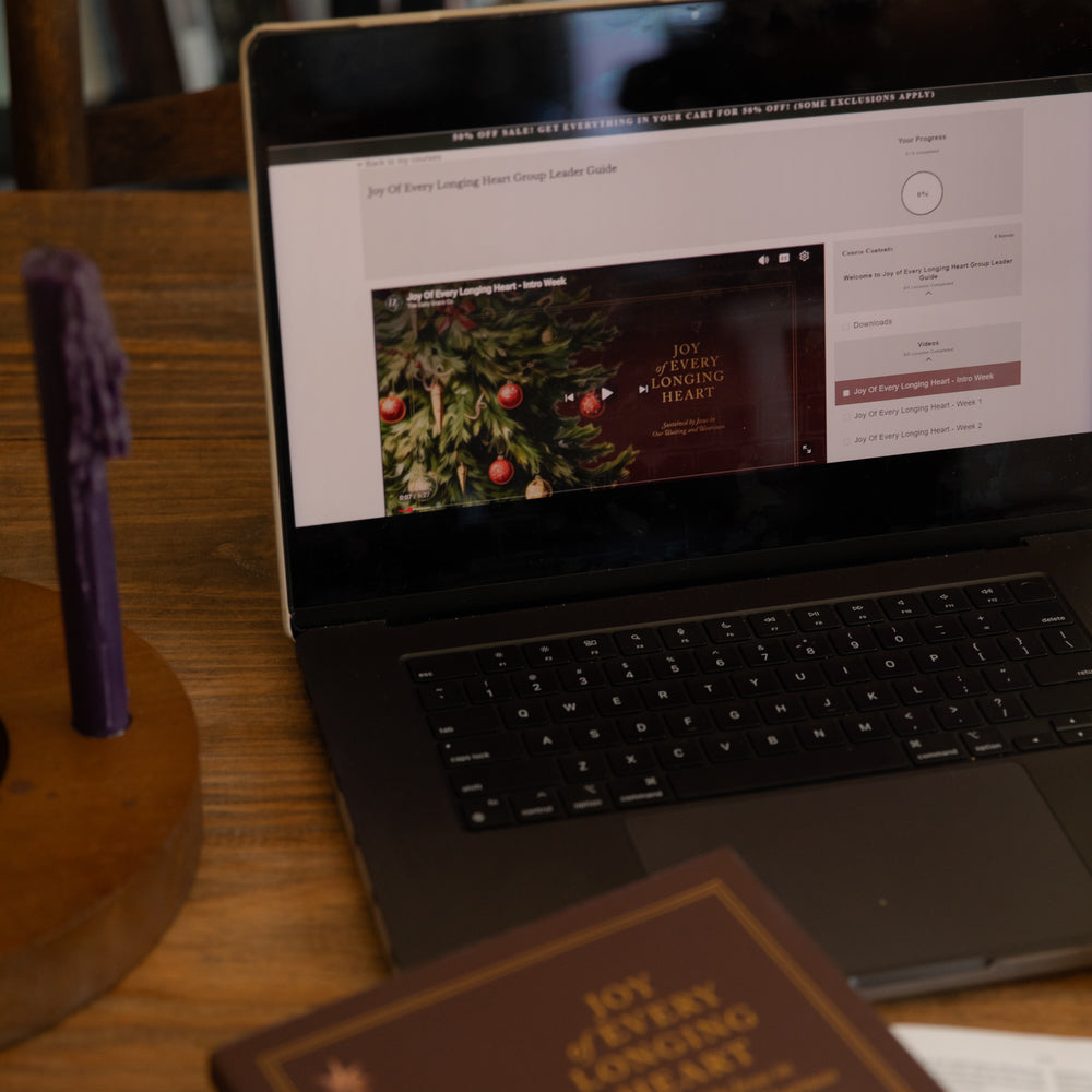 A laptop on a wooden table displaying a website template or digital guide, with a purple candle in a wooden holder on the left and a brown book in the foreground.