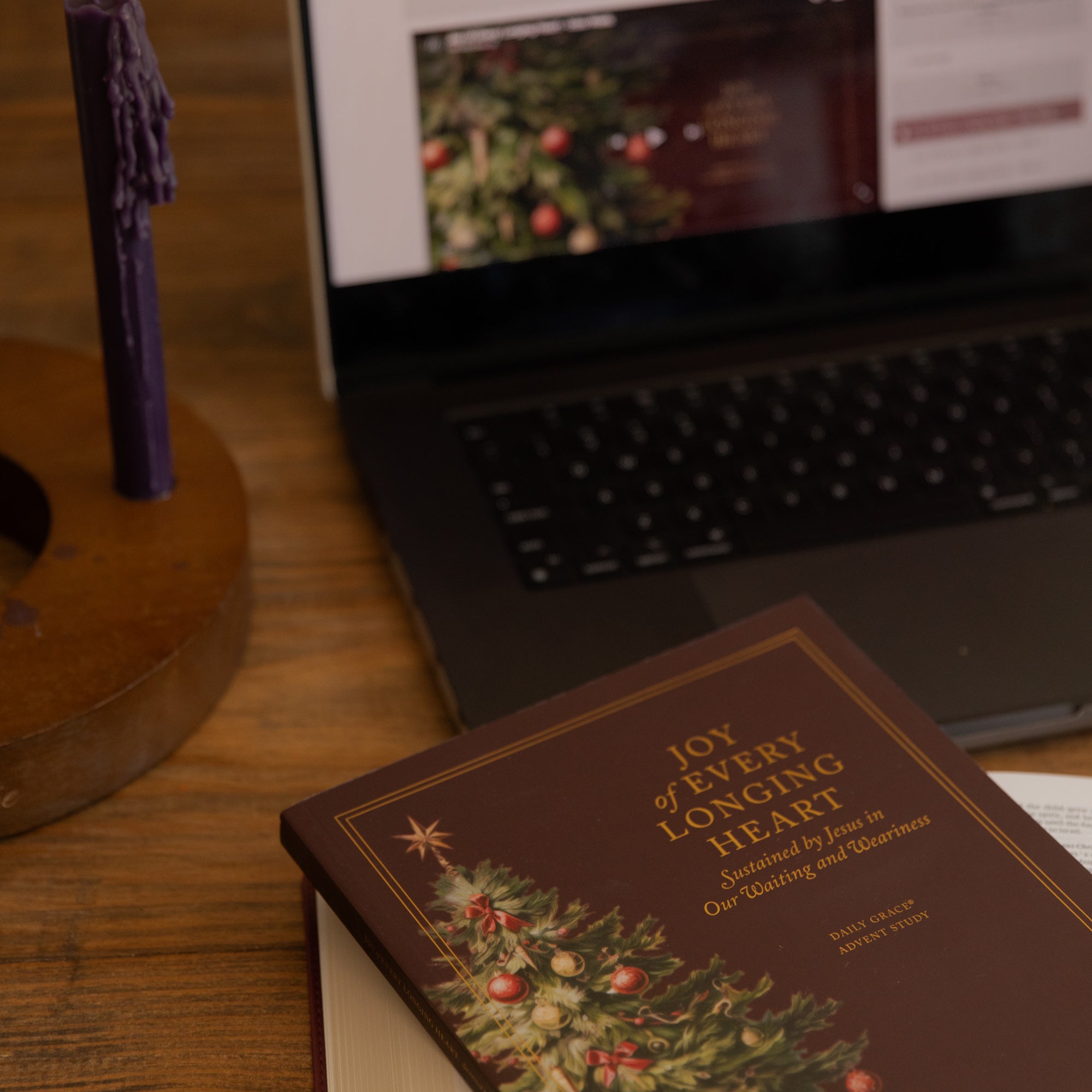 A brown book titled 'Joy of Every Longing Heart: Sustained by Jesus in Our Waiting and Weariness' with a Christmas tree illustration on the cover, resting on a wooden table next to an open laptop and a purple candle.