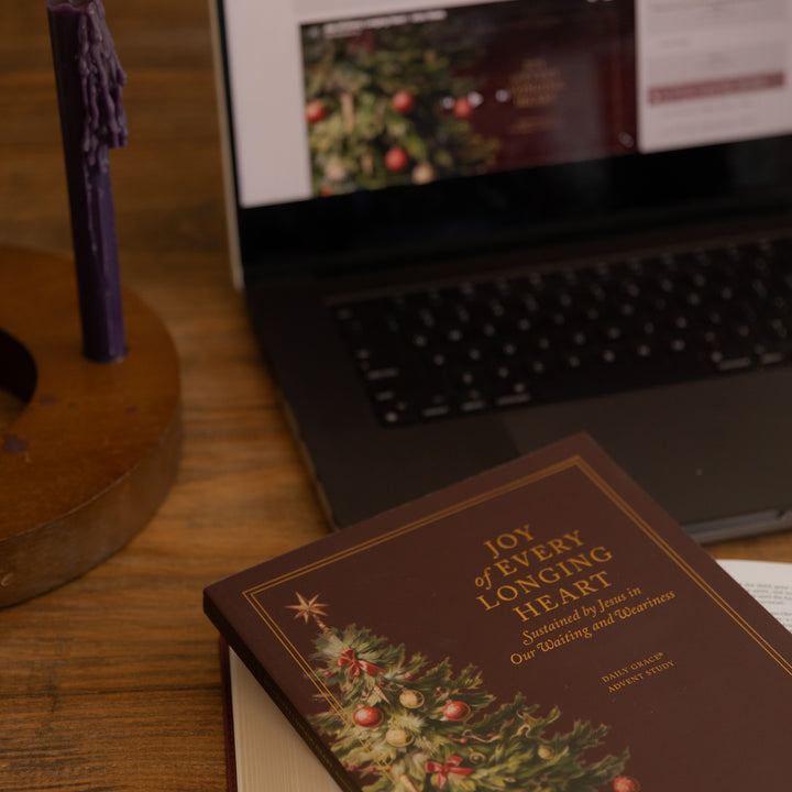 A brown book titled 'Joy of Every Longing Heart: Encountering Joy in Our Scattered and Sometimes Broken Heart' with a Christmas tree illustration on the cover, resting on a wooden table next to an open laptop and a purple candle.