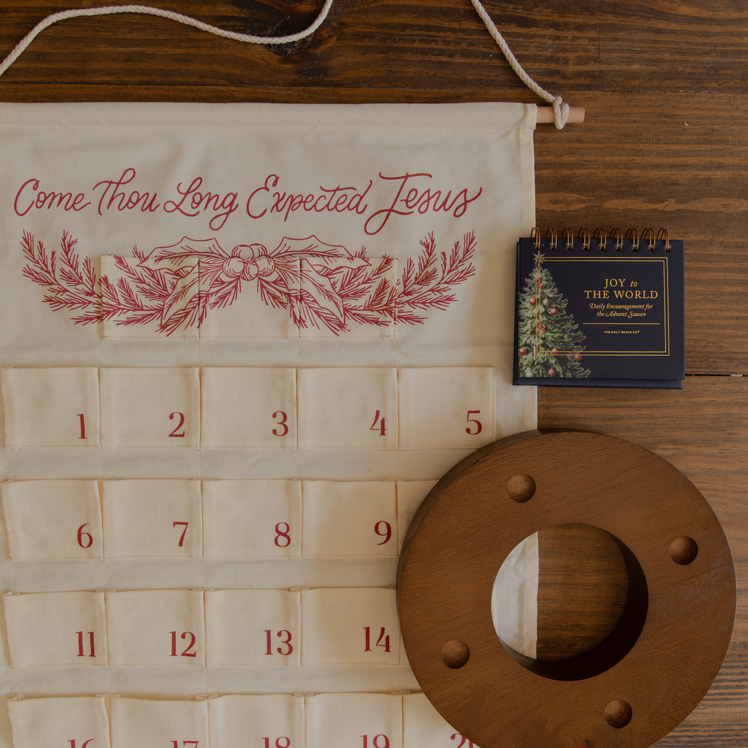An Advent calendar made from cream-colored fabric with red embroidered text reading 'Come Thou Long Expected Jesus' and red number tabs, laid flat on a dark wood surface alongside a small spiral notebook and a round wooden dowel.