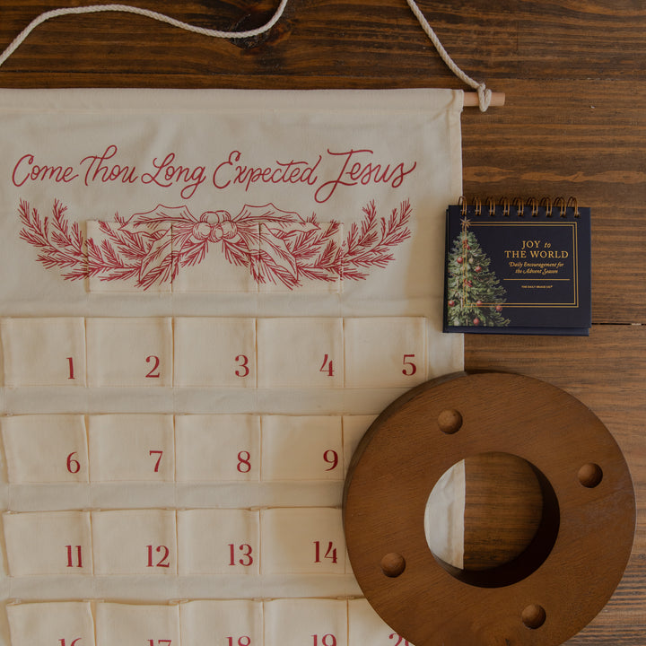 An Advent calendar made from cream-colored fabric with red embroidered text reading 'Come Thou Long Expected Jesus' and red number tabs, laid flat on a dark wood surface alongside a small spiral flip book and a round wooden dowel.