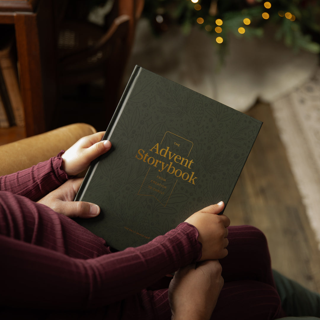 A person holding a dark green hardcover book titled 'The Advent Storybook' with gold lettering, shown in a cozy indoor setting near a Christmas tree with warm lights.