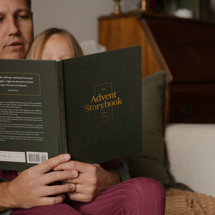 A man and a child are sitting together indoors, reading a dark green hardcover book titled "The Advent Storybook" with gold lettering. The scene is softly lit and includes pillows and a wooden cabinet in the background.