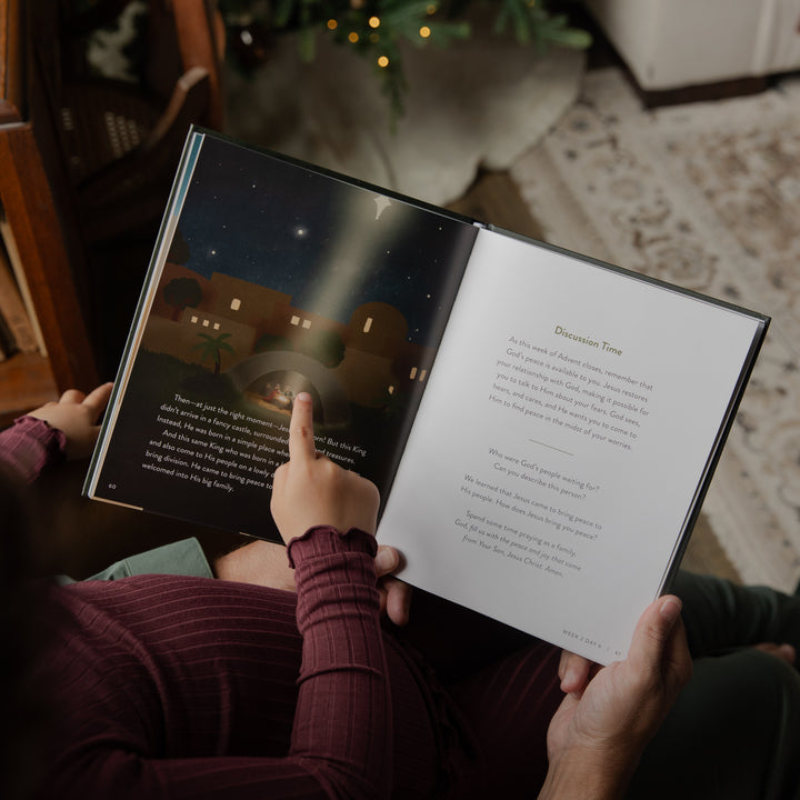 A person holding an open children's book in a cozy indoor setting, pointing at a starry night scene on one page while reading text on the other page.