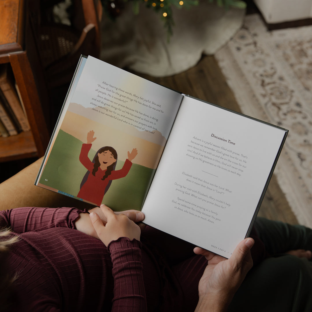 A person sitting on a couch holds an open children's book with illustrated and text pages, in a cozy indoor setting with a rug and furniture visible.
