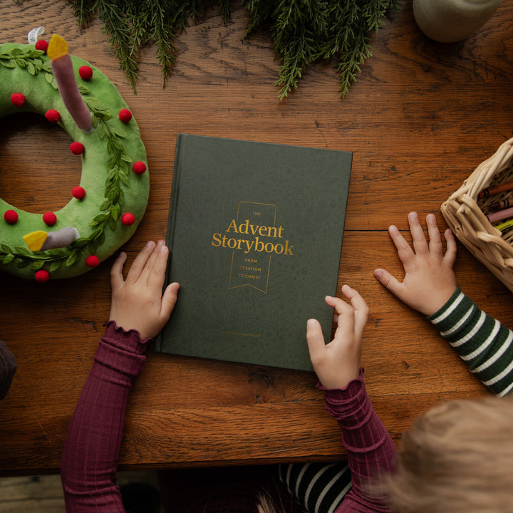 A child's hands hold a dark green hardcover book titled 'The Advent Storybook' on a rustic wooden table, surrounded by holiday decorations including a green wreath, evergreen branches, and a basket of crayons.