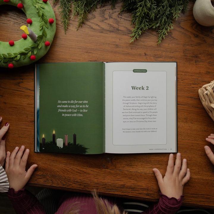 An open book laid on a wooden table, showing a green left page with a Bible verse and a white right page with text titled 'Week 2.' Hands rest on the table around the book, with holiday greenery and a wreath visible in the scene.