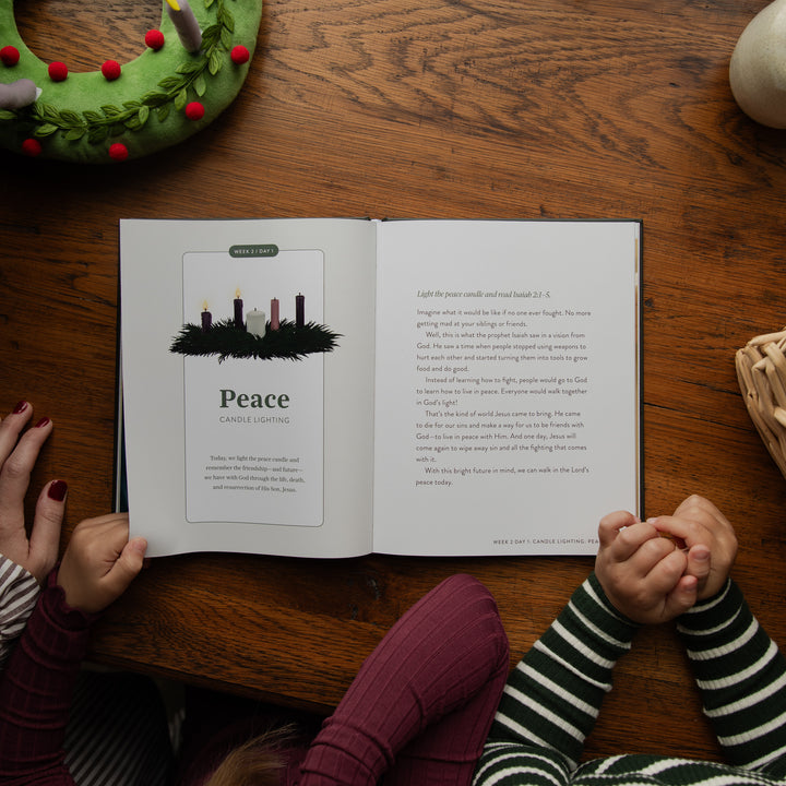 An open children's book titled 'Peace' is laid on a wooden table, viewed from above. Two children's hands and arms in striped sweaters are visible holding the book open.