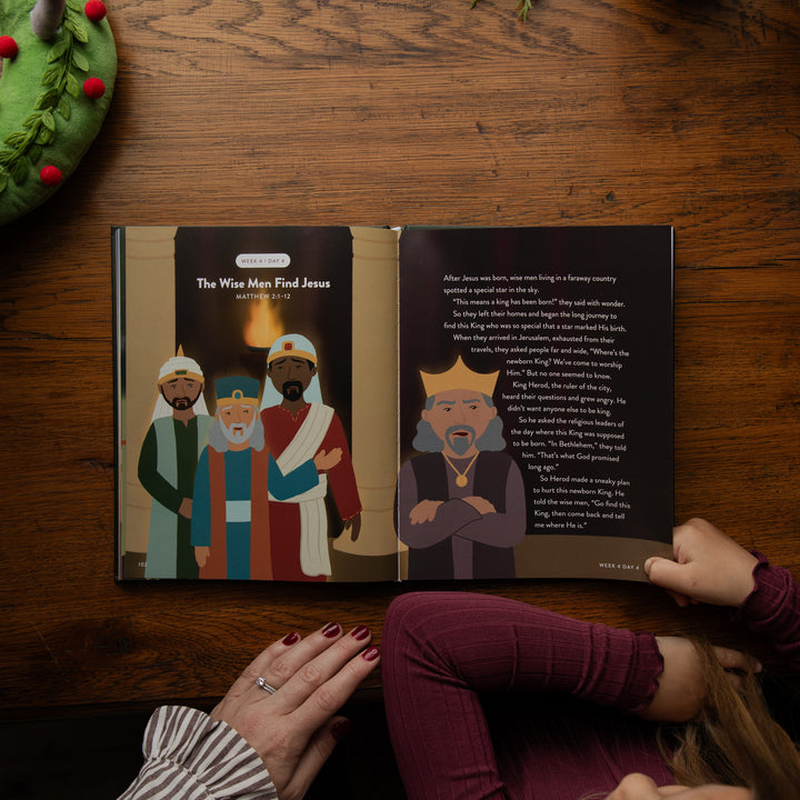 An open children's Bible or storybook spread on a wooden table, showing illustrated figures and text about King Herod and the Wise Men. Two children's hands are visible holding or resting near the book, and part of a green wreath appears in the top left corner.
