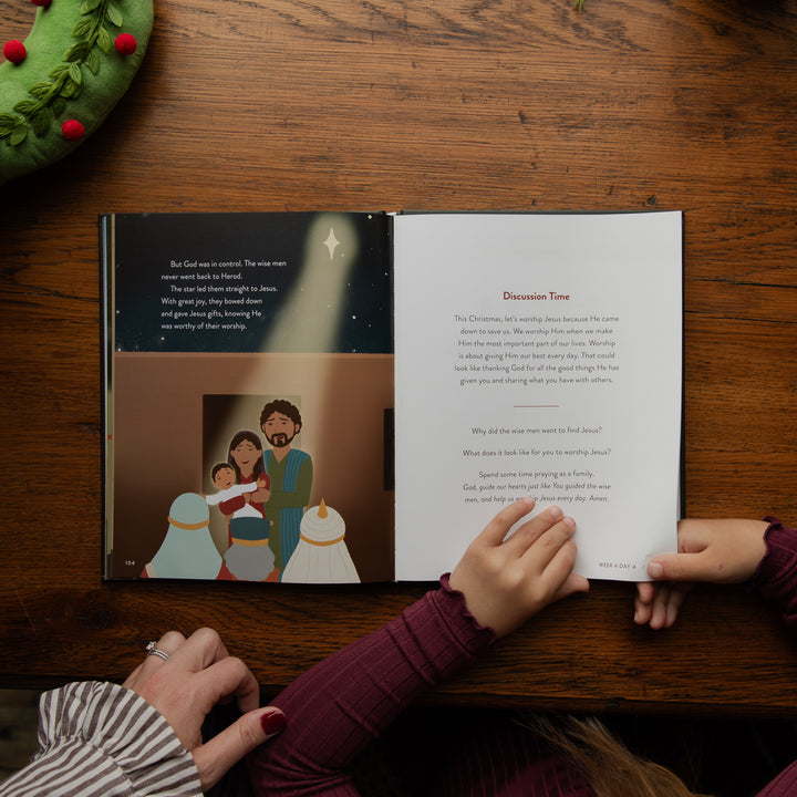 An open children's Bible or scripture book laid on a wooden table, with two children's hands holding the pages and another hand resting nearby. A green wreath is partially visible in the top left corner.