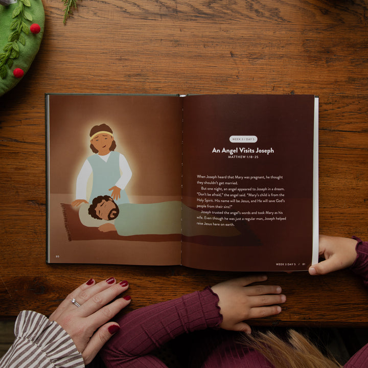 An open children's Bible or storybook spread on a wooden table, showing an illustration of an angel visiting Joseph and a Bible verse from Matthew 18:14. Hands are visible holding and resting on the book, with a Christmas wreath partially visible in the top left corner.
