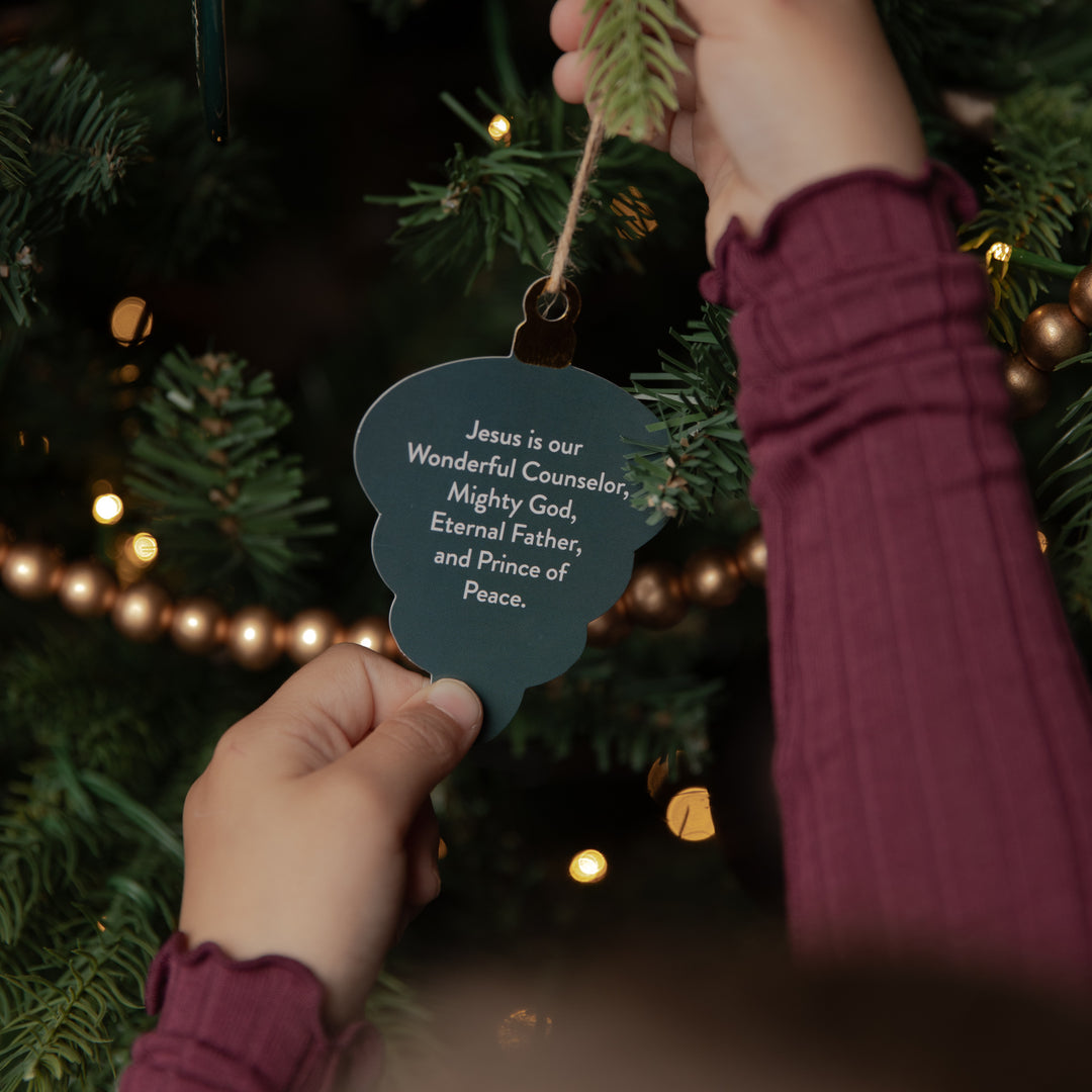 A person hanging a dark teal cloud-shaped ornament on a Christmas tree. The ornament has white printed text that reads, 'Jesus is our Wonderful Counselor, Mighty God, Eternal Father, and Prince of Peace.