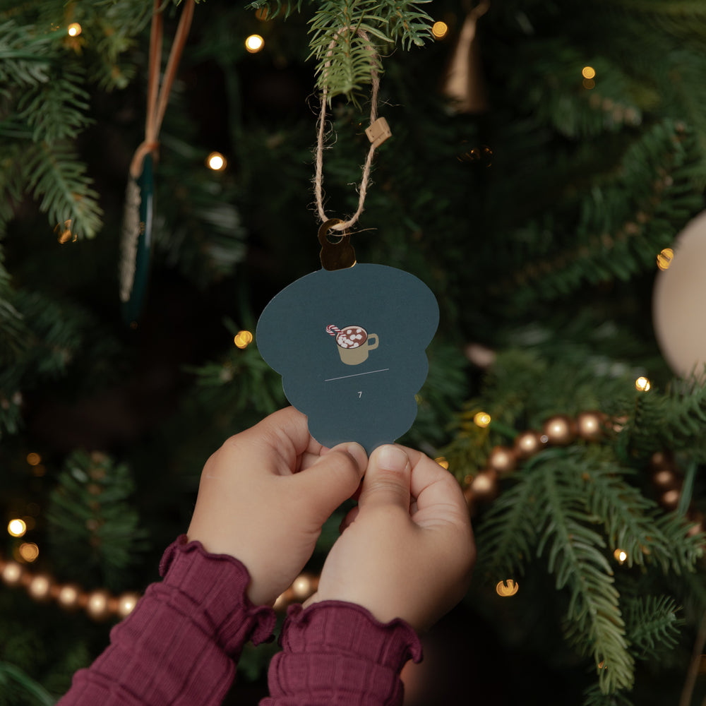 A person's hands wearing a burgundy long-sleeve top hang a dark teal Christmas ornament shaped like a mug on a decorated Christmas tree with warm string lights.
