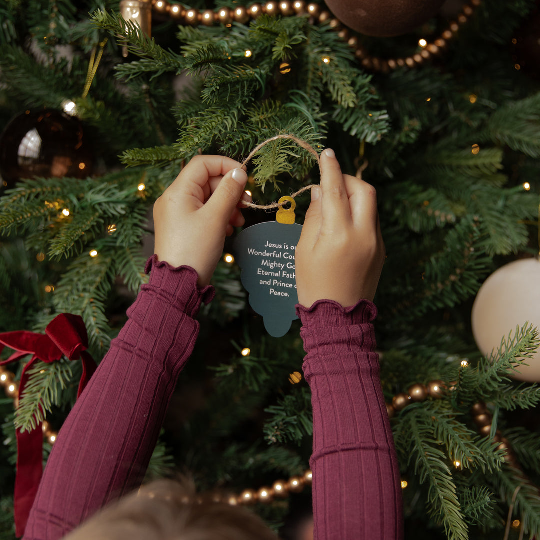 A person wearing a burgundy ribbed long-sleeve top hangs a small cloud-shaped ornament on a decorated Christmas tree. The ornament has a light brown twine loop and a small yellow tag, with white text that reads, 'Jesus is the Wonderful Savior, Mighty God, Eternal Father, and Redeemer, Peace.