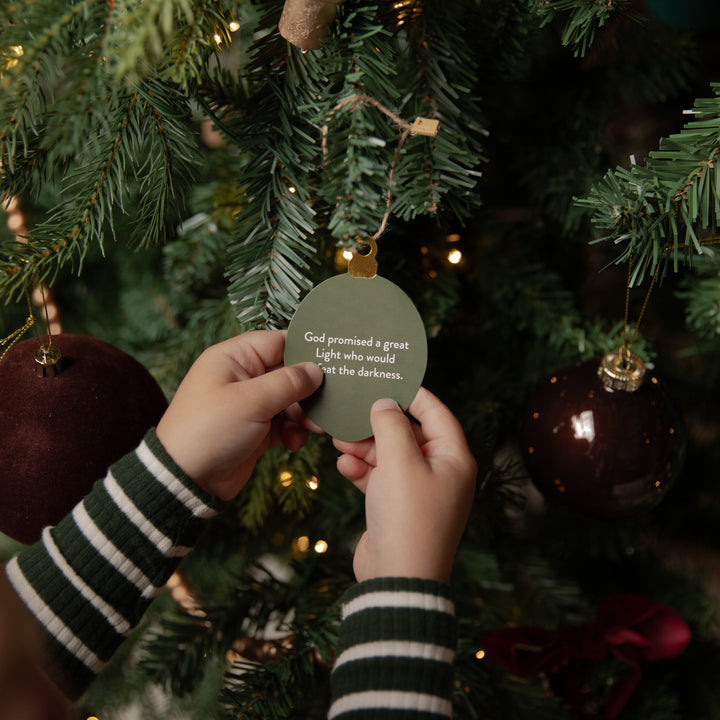 A person's hands wearing green and white striped sleeves hang a round sage green ornament on a Christmas tree. The ornament features the printed quote, 'God promised a great Light who would shine out the darkness.