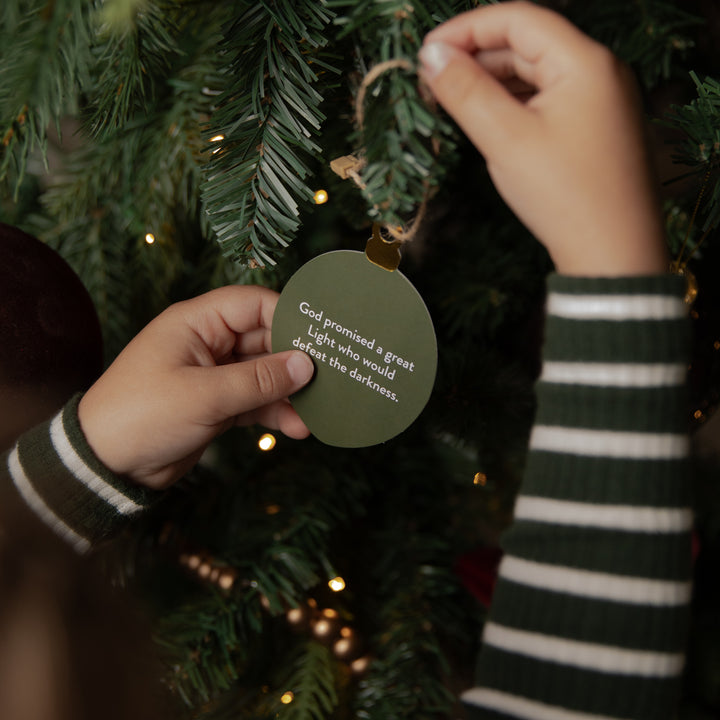 A person hanging a round olive green Christmas ornament on a decorated Christmas tree. The ornament has white printed text that reads, 'God promised a great light who would dazzle the darkness.