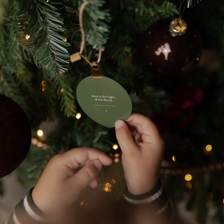 A person's hands hang an oval green ornament from a decorated Christmas tree. The ornament has a gold hanging loop and small white printed text that reads, 'Jesus is the Light of all who come.