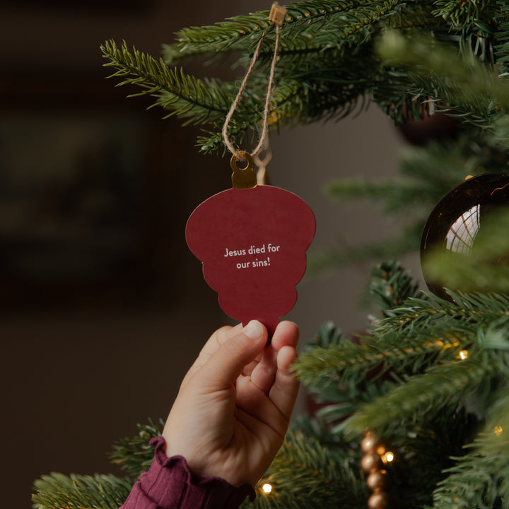 A hand hangs a red Christmas ornament shaped like a light bulb from a green Christmas tree. The ornament has white text that reads 'Jesus died for our sins!' and is suspended by a twine string with a gold-colored metal loop.