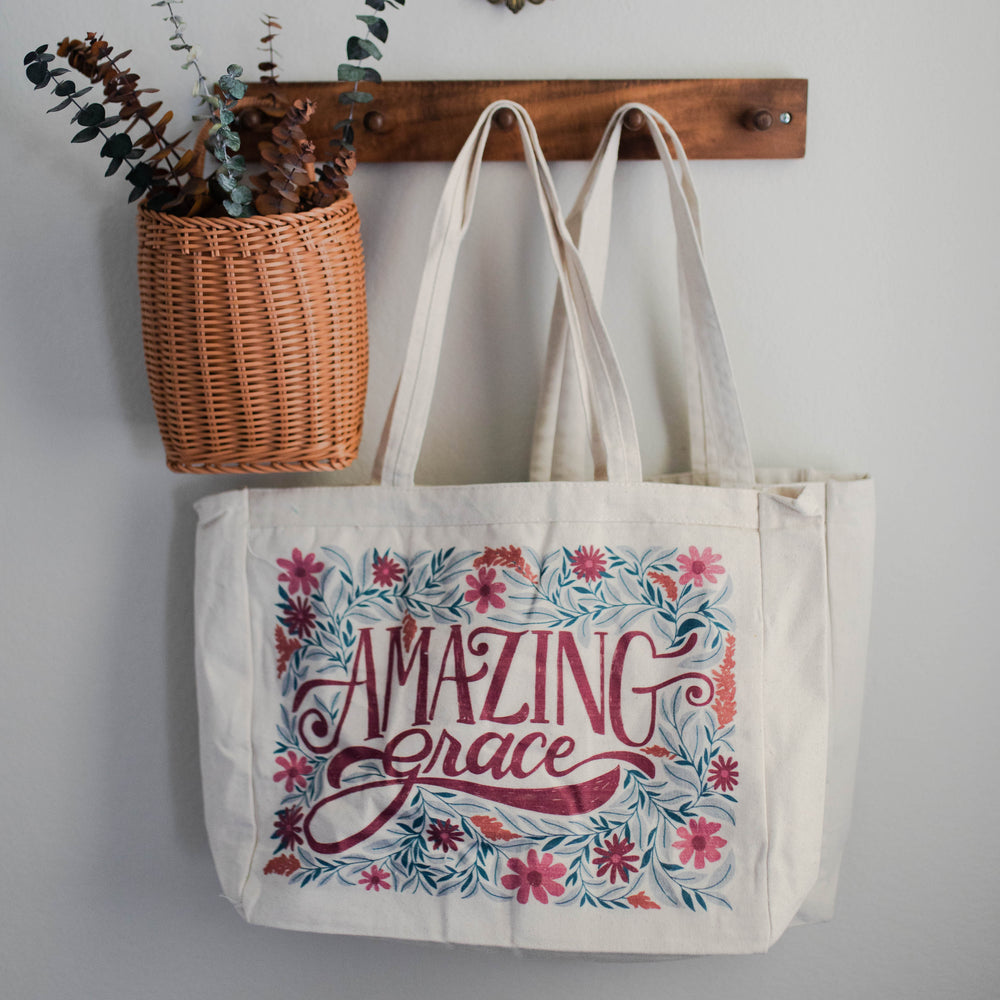 Printed tote that reads "amazing grace" with colorful florals hanging on rack