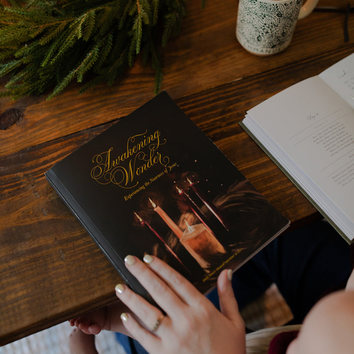 The Awakening Wonder Cover on a wooden table with a woman's hand resting on the corner near some greenery and a coffee cup