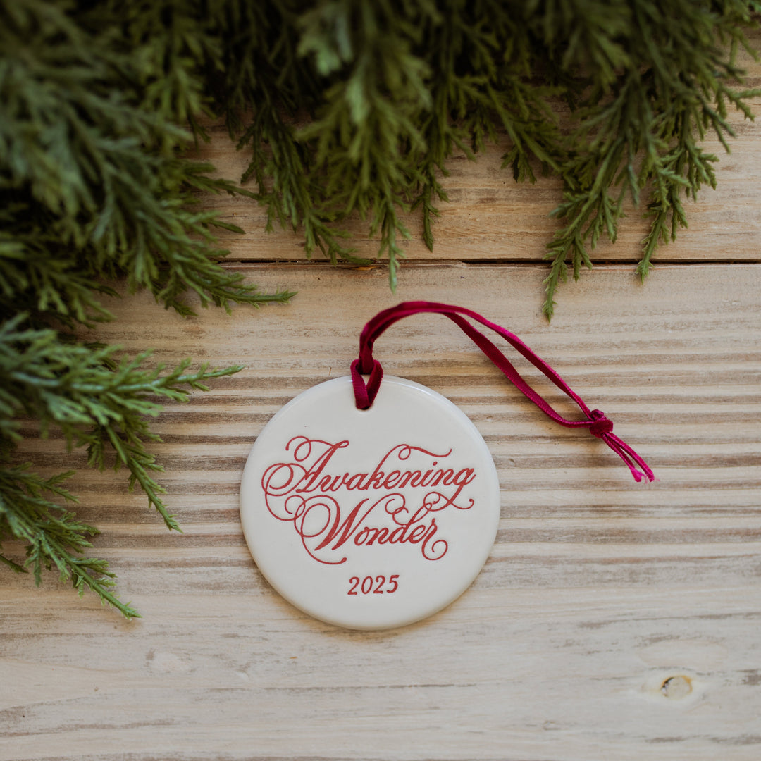 Round ornament with 'Awakening Wonder 2025' text on a wooden surface with greenery