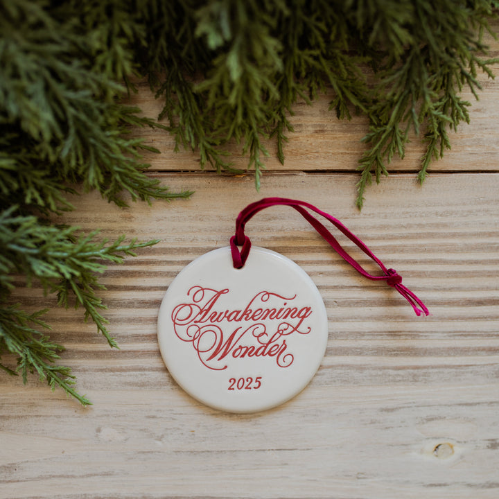 Round ornament with 'Awakening Wonder 2025' text on a wooden surface with greenery