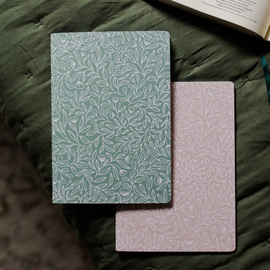 Two stitched journals with floral designs laying on a couch with a Bible in the background. 