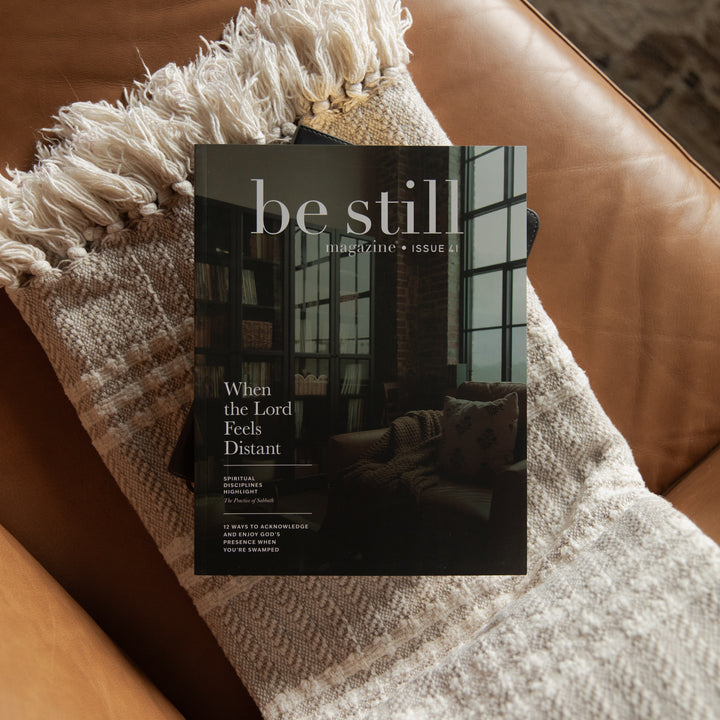 A magazine titled "be still" placed on a brown leather couch, resting on a cream woven throw blanket with fringe.