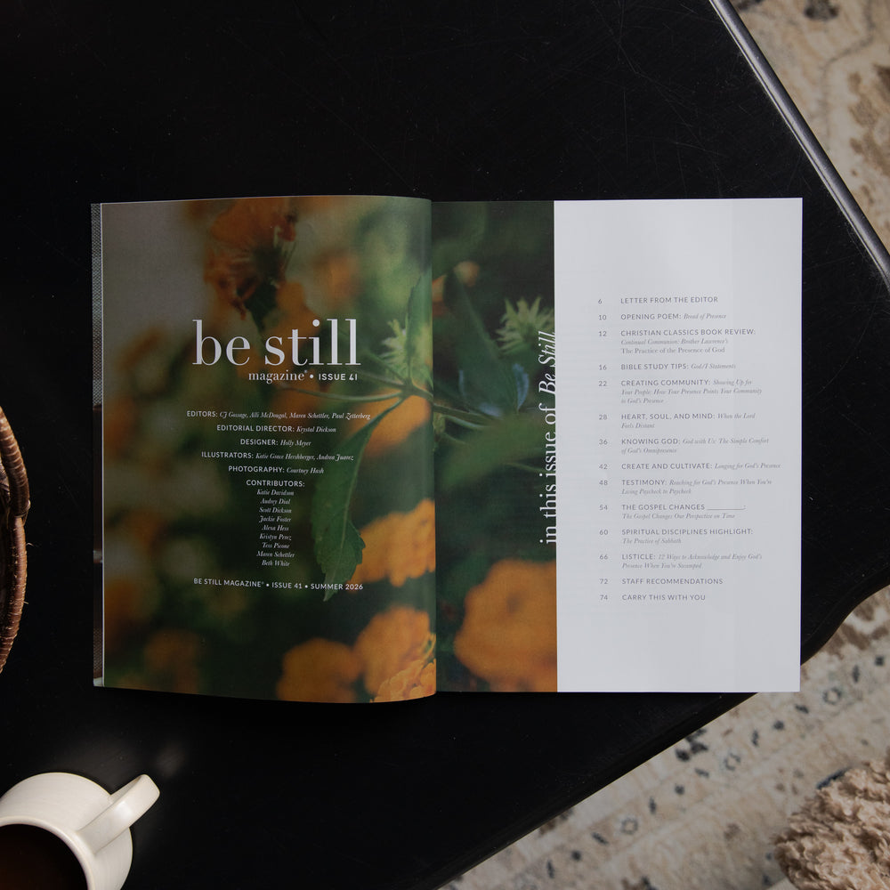 An open magazine or book titled "be still" lies on a dark tabletop, showing a floral cover spread with a white page of text on the right. A white cup is partially visible in the lower left corner.