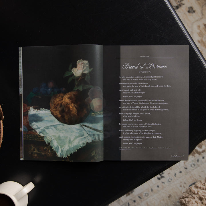 An open printed book or magazine laid on a dark surface, showing a still life painting on the left page and a poem titled 'Bread of Presence' on the right page.